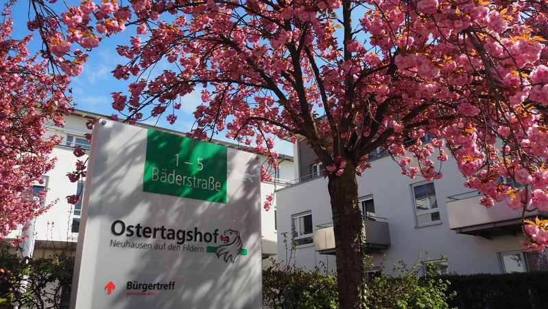 Bürgertreff Ostertagshof: Ein blühender Baum steht neben einem Schild mit der Aufschrift "1-5 Bäderstraße, Ostertagshof" vor einem weißen Gebäude.