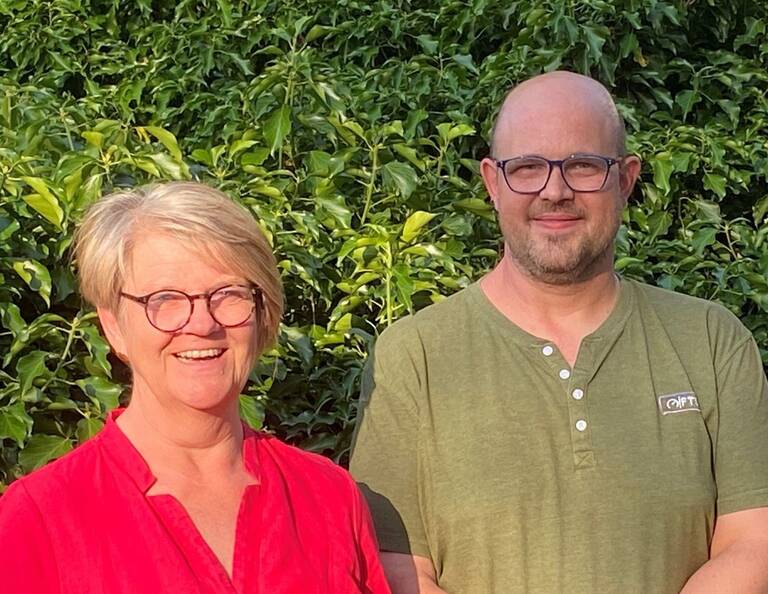 Eine lächelnde Frau in einem roten Oberteil und ein Mann in einem grünen T-Shirt stehen vor einer grünen Blatthecke.
